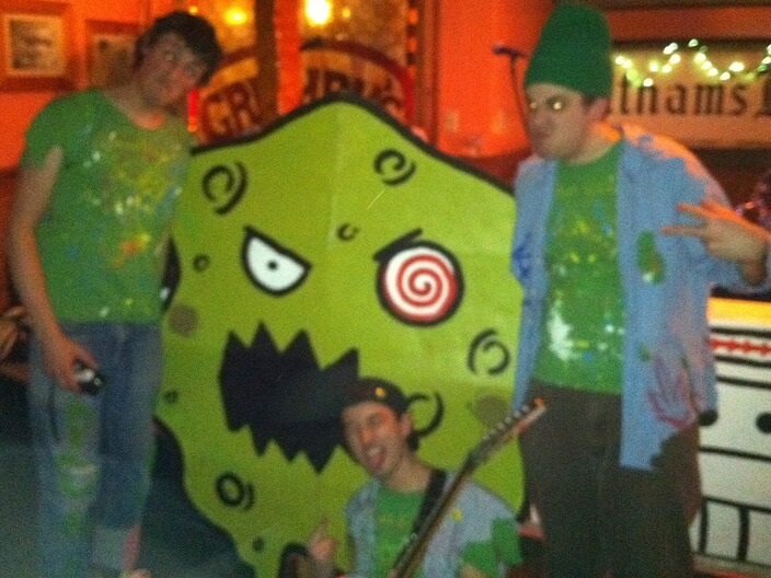 Three paint-splattered weirdos pose in front of a human-sized amboeba for their really cool-looking band photo.