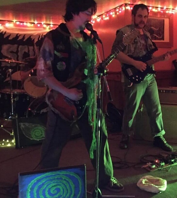 Lord Scum performing live. Algaenon closes his eyes while incanting into the microphone, guitar in hand. Oozerus gazes directly into our souls, looking cool in his denim vest.